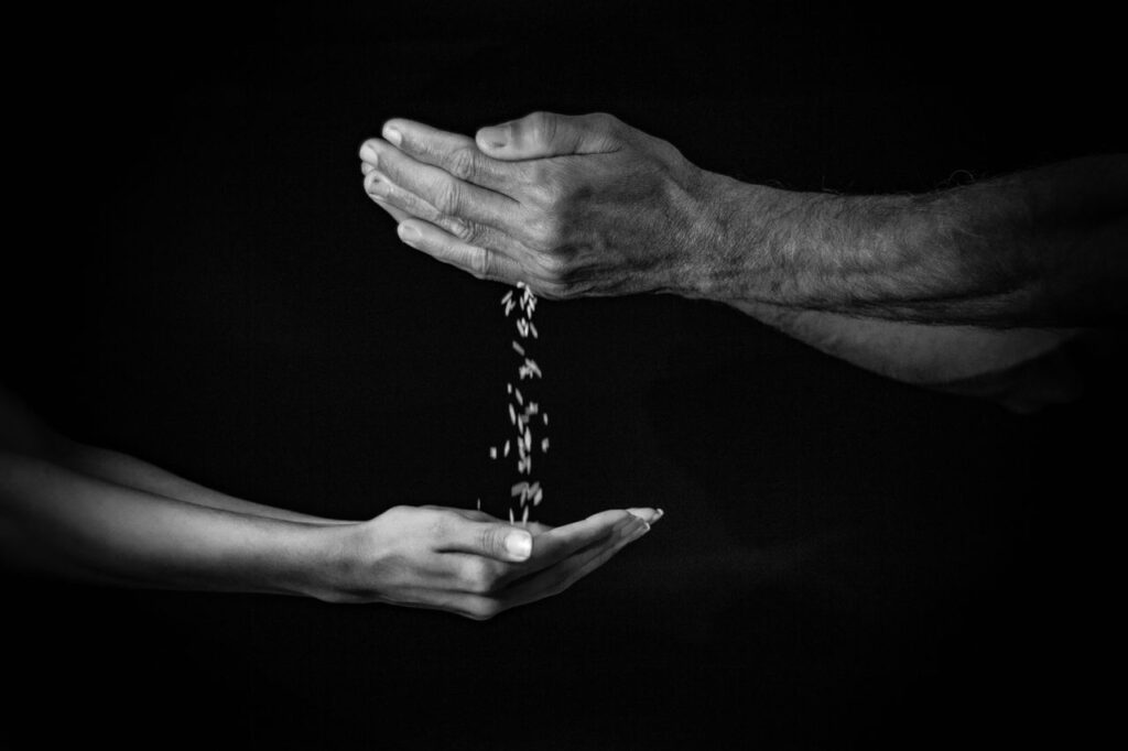 Black and white image of hands exchanging falling grains symbolizing charity or passage of time.