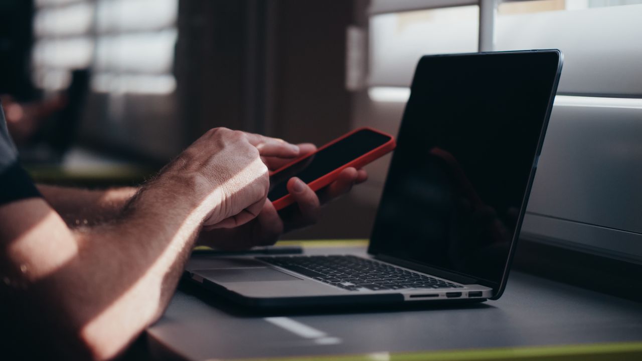 A person holding a smartphone near a laptop in an indoor setting, depicting modern technology use.